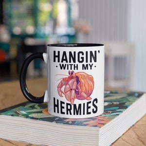 May include: White ceramic mug with a black rim and handle. The mug has a black and white graphic of a hermit crab with the text "Hangin' with my Hermies".