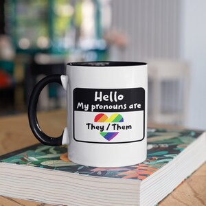 Op de afbeelding: Witte keramische mok met een zwarte handgreep en binnenkant. De mok heeft een labelontwerp met de tekst "Hello, My pronouns are They/Them" met een regenbooghart. De mok staat op een stapel boeken.