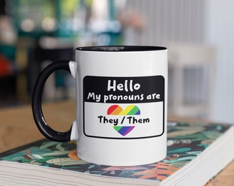 May include: White ceramic mug with a black handle and interior. The mug features a label design that reads "Hello, My pronouns are They/Them" with a rainbow heart. The mug is sitting on a stack of books.