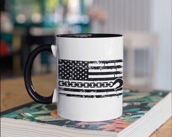 May include: White ceramic mug with a black handle and interior. The mug features a distressed American flag design with a chain and hook graphic in black and white. The mug is sitting on a book.