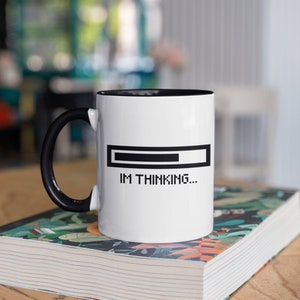 May include: White ceramic mug with a black interior and handle. The mug features a black loading bar graphic and the text "IM THINKING...". The mug is sitting on a book with a floral design.