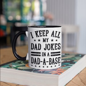 May include: White coffee mug with a black handle and black rim. The mug has a black graphic that says "I keep all my dad jokes in a dad-a-base" with stars around the text.