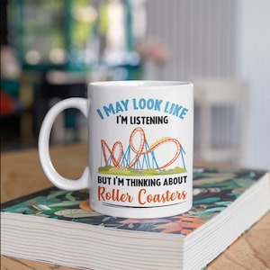 Könnte beinhalten: Weiße Keramik-Tasse mit dem Text "I MAY LOOK LIKE I'M LISTENING BUT I'M THINKING ABOUT Roller Coasters" und einer Achterbahn-Grafik. Die Tasse steht auf einem Buch mit floralem Design.