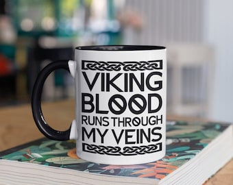 May include: White ceramic mug with a black handle and interior. The mug features the text "VIKING BLOOD RUNS THROUGH MY VEINS" in black, with decorative knotwork. The mug is sitting on a book with a floral design.