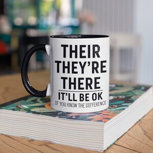Puede incluir: Taza de cerámica blanca con asa e interior negros, con el texto "THEIR THEY'RE THERE IT'LL BE OK IF YOU KNOW THE DIFFERENCE" en letras negras. La taza está sobre un libro con una cubierta floral.