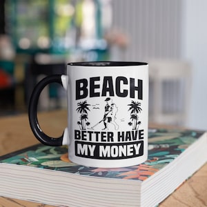May include: White ceramic mug with a black handle and interior. The mug features the text "BEACH BETTER HAVE MY MONEY" in black, along with a graphic of a person metal detecting on a beach, and palm trees.
