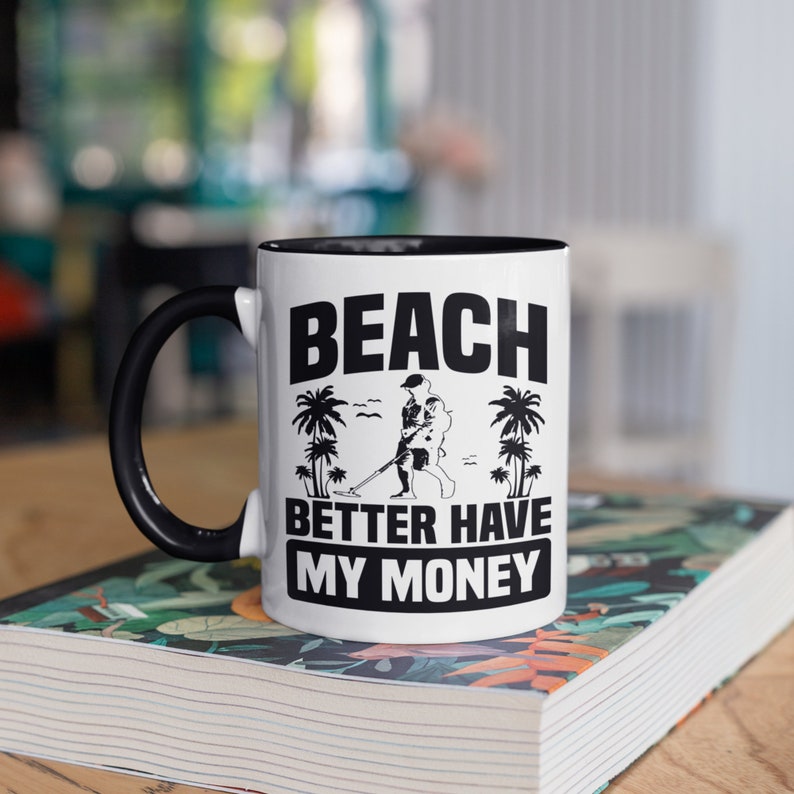 May include: White ceramic mug with a black handle and interior. The mug features the text "BEACH BETTER HAVE MY MONEY" in black, along with a graphic of a person metal detecting on a beach, and palm trees.