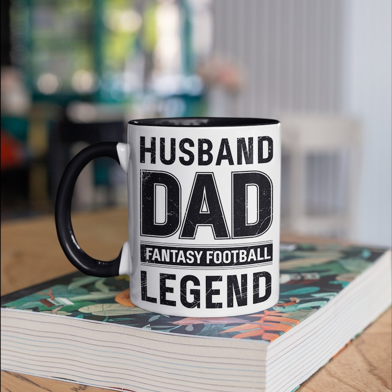 May include: White ceramic mug with a black handle and interior. The mug features the words "HUSBAND DAD FANTASY FOOTBALL LEGEND" in a bold, distressed font. The mug is sitting on a book with a floral design.