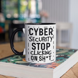 Op de afbeelding: Witte keramische mok met een zwarte handgreep en binnenkant. De mok heeft de tekst "CYBER SECURITY STOP CLICKING ON SH*T" in zwart, met een afspeelknop. De mok staat op een boek met een kleurrijk bloemenontwerp.