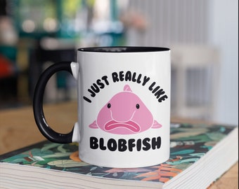 May include: White ceramic mug with a black handle and interior. The mug features a pink blobfish illustration and the text "I JUST REALLY LIKE BLOBFISH" in black. The mug is sitting on a book.