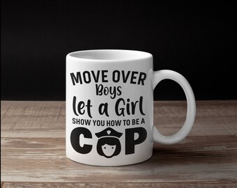 May include: White ceramic mug with the text "MOVE OVER Boys let a Girl SHOW YOU HOW TO BE A COP" in black. The mug features a cartoon illustration of a police officer. The mug is on a wooden surface.