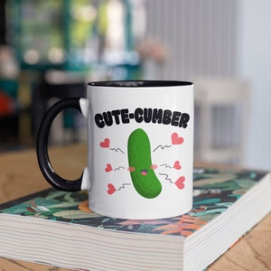 May include: White ceramic mug with a black handle and interior. The mug features the text "CUTE-CUMBER" above a cartoon pickle with hearts and wavy lines. The mug is sitting on a patterned book.