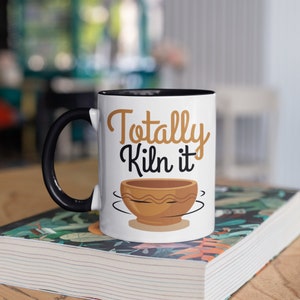May include: White ceramic mug with a black rim and handle. The mug has a brown and yellow illustration of a pottery bowl and the text "Totally Kiln it".