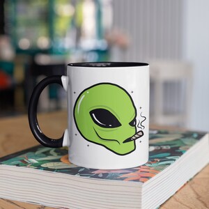 Op de afbeelding: Witte keramische mok met een zwarte handgreep en binnenkant. De mok heeft een groene alienkop met zwarte ogen en een brandende sigaret. De mok staat op een boek met een bloemenmotief.