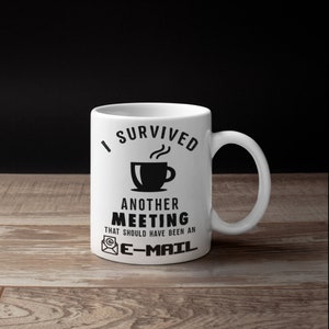 May include: White ceramic coffee mug with the text "I SURVIVED ANOTHER MEETING THAT SHOULD HAVE BEEN AN E-MAIL" in black. The mug features a black coffee cup graphic with steam rising from it. The mug has a curved handle.