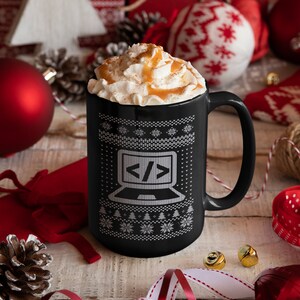 May include: A black coffee mug with a white knit pattern and a laptop graphic with code symbols. The mug is filled with whipped cream and caramel drizzle. Festive holiday decorations surround the mug, including ornaments and ribbons.