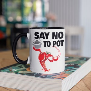 May include: White ceramic mug with a black handle and interior. The mug features the text "SAY NO TO POT" above a cartoon crawfish holding a pot. The mug is sitting on a book with a floral design.