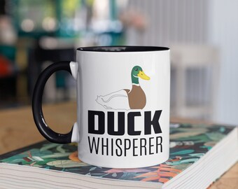 Puede incluir: Taza de cerámica blanca con asa e interior negros. La taza presenta una ilustración de un pato de dibujos animados y las palabras "DUCK WHISPERER" en letras negras en negrita. La taza está sobre un libro con diseño floral.