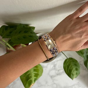 May include: A rectangular wristwatch with a gold-tone case and a silver and purple band is worn on a wrist. A gold bracelet and a black leather band are also visible. The watch face is a light colour. Green leaves are in the background.