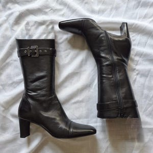Puede incluir: Un par de botas negras de media caña con punta cuadrada, tacón de bloque y una tira decorativa con hebilla de metal plateado en la parte superior. Las botas tienen un acabado liso, una cremallera lateral vertical y una costura en la puntera.
