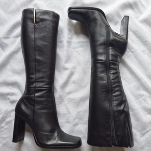May include: A pair of black knee-high boots featuring a square toe design and a sturdy block heel. The boots are crafted from smooth black leather and include a full-length side zipper closure for easy wear, with a clean, structured silhouette.