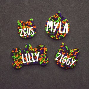 May include: Four colorful pet ID tags with black, green, and orange glitter. The tags are shaped like a bone, a cat, a circle, and a cat. The names "Lilly", "Zeus", "Myla", and "Ziggy" are written in white on the tags.