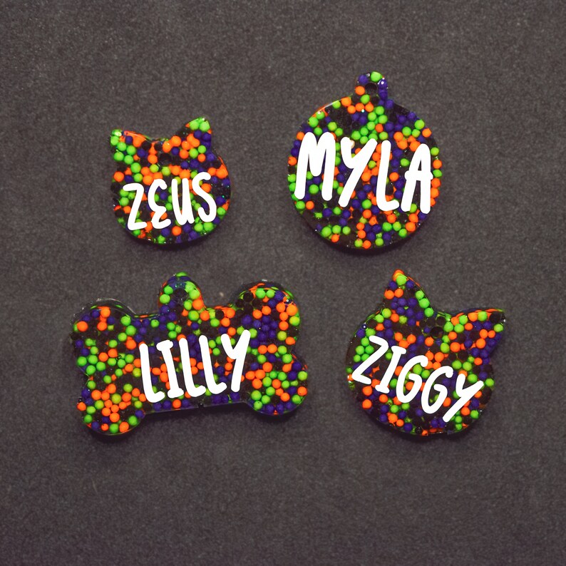 May include: Four colorful pet ID tags with black, green, and orange glitter. The tags are shaped like a bone, a cat, a circle, and a cat. The names "Lilly", "Zeus", "Myla", and "Ziggy" are written in white on the tags.