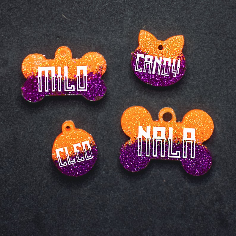 May include: Four orange and purple glitter dog tags with white text. The tags are shaped like a bone, a cat, and two circles. The text on the tags reads "Milo", "Candy", "Nola", and "Cleo".