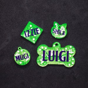 May include: Four green pet ID tags with white ghost designs and purple lettering. The tags are shaped like a bone, a diamond, a circle, and a cat head. The names on the tags are "Luigi", "Izzie", "Mugi", and "Shila".