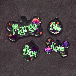 May include: Four Halloween-themed dog tags with glitter and pumpkin accents. The tags read "Margo", "Boo", "Koko", and "Binx".