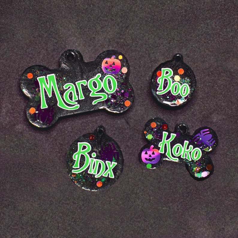 May include: Four Halloween-themed dog tags with glitter and pumpkin accents. The tags read "Margo", "Boo", "Koko", and "Binx".