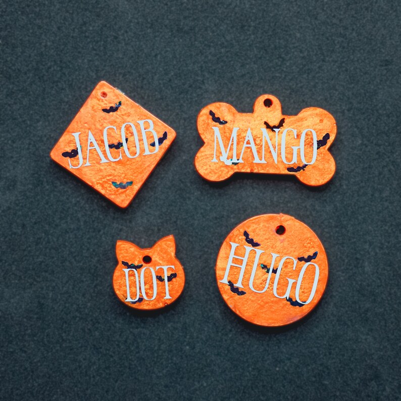 May include: Four orange Halloween-themed pet ID tags with black bats and white lettering. The tags read "Jacob", "Mango", "Hugo", and "Dot".