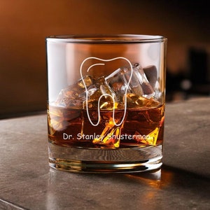 May include: A clear glass tumbler with a tooth design and the text "Dr. Stanley Shusterman" etched on the side. The glass is filled with amber liquid and ice cubes.