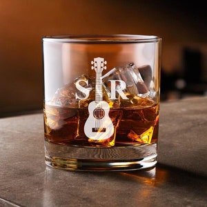 May include: A clear glass rocks glass with a white etched design of a ukulele and the letters "SR". The glass is filled with amber liquid and ice cubes.