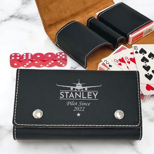 May include: Black leather card and dice case with a white airplane graphic and the text "STANLEY Pilot Since 2022". The case has a brown suede interior and is open to reveal three card slots and a deck of cards.