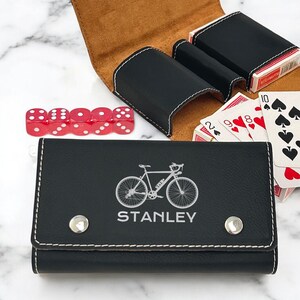 May include: Black leather card and dice case with a white bicycle design and the name "STANLEY" engraved on the front. The case has two slots for decks of cards and a small compartment for dice.
