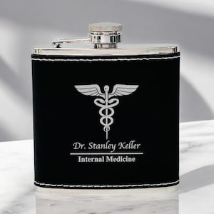 May include: Black leather flask with a silver cap and a silver medical symbol engraved on the front. The text "Dr. Stanley Keller Internal Medicine" is also engraved on the flask.