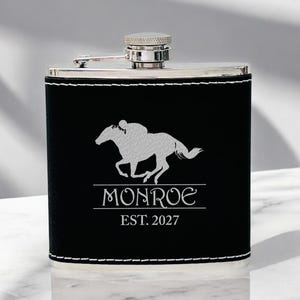 May include: Black leather flask with a silver top and a silver horse and rider silhouette. The flask is engraved with the text "Monroe Est. 2027".