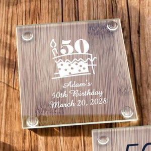 May include: Two square glass coasters with a wood-grain design. The top coaster features a white cake with a candle and the text "50 Adam's 50th Birthday March 20, 2028". The bottom coaster features a black and white design with the text "50th Birthday Bash! Albert's 50th Birthday June 11, 2027".