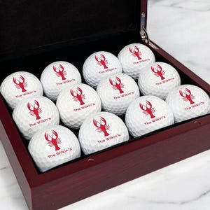 May include: A mahogany wood box with a hinged lid, containing twelve white golf balls with a red lobster logo and the text "The Wicker's" printed on each ball.