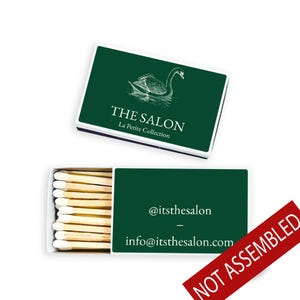 May include: A matchbox with a dark green cover featuring a swan illustration and the text "THE SALON La Petite Collection." The open box reveals matchsticks and a dark green interior with the text "@itsthesalon" and "info@itsthesalon.com."