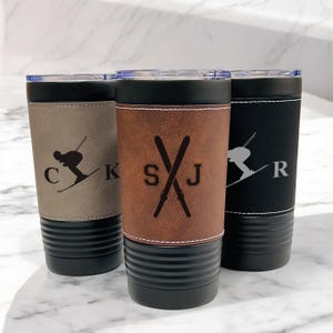 May include: Three personalized insulated tumblers with black bases and lids. The tumblers have a leather-like wrap with a skier graphic and initials. One is tan, one is brown, and one is black. The initials are "C", "KSJ", and "R".