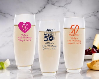 May include: Three champagne flutes with personalized 50th birthday messages. The first flute has a pink heart with the number 50 inside and the text "Julia's 50th Birthday May 4, 2026". The second flute has the text "You Made The Big 50" with a blue and white illustration of a fishing rod and fish. The text "Albert's 50th Birthday June 11, 2027" is below the illustration. The third flute has the number 50 in orange with the text "and fierce! Kelly's 50th Birthday April 5, 2029".