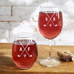 May include: Two wine glasses with personalized golf-themed designs. The larger glass has "SR" with crossed golf clubs and a golf ball. The smaller glass has "AF" with crossed golf clubs and a golf ball.