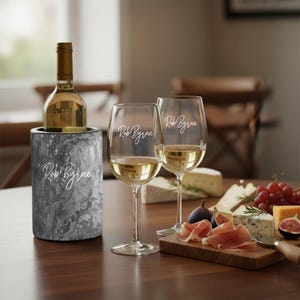 May include: A wine bottle in a marble-patterned cooler, two wine glasses, and a wooden board with cheese, grapes, and figs. The wine glasses and cooler have the text "Rob Byrne" in a script font. The wine glasses are filled with white wine.