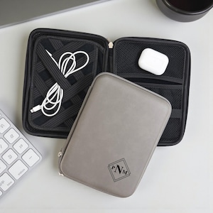 May include: Gray zippered tech organizer with a monogram, open to reveal a white charging cable and a black mesh pocket with white earbuds. The organizer is on a white surface.