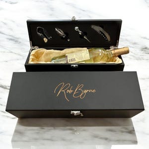 May include: A black rectangular wooden wine gift box with a metal latch, featuring the name Rob Byrne in gold script on the lid. The open box contains a clear glass wine bottle nestled in beige fabric, alongside four silver wine accessories.