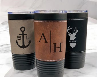 May include: Three stainless steel tumblers with different designs. The first tumbler has a gray leather wrap with an anchor and the initials "S.L." The second tumbler has a brown leather wrap with the initials "A.H." The third tumbler has a black leather wrap with a deer silhouette and the name "STEVE".