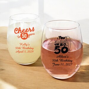 May include: Two clear wine glasses with personalized text for a 50th birthday. The glass on the left has the text "Cheers to 50 years" with the number 50 in orange. The glass on the right has the text "You Made the Big 50!" with the number 50 in black. Both glasses have the name of the birthday person and the date of the birthday.