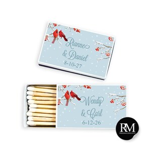 May include: Two rectangular matchboxes with a light blue winter background featuring red cardinals on snowy branches with red berries. One box is closed, the other is open showing wooden matches. Text reads Rianne & Daniel 8-10-27 and Wendy & Carl 6-12-26.